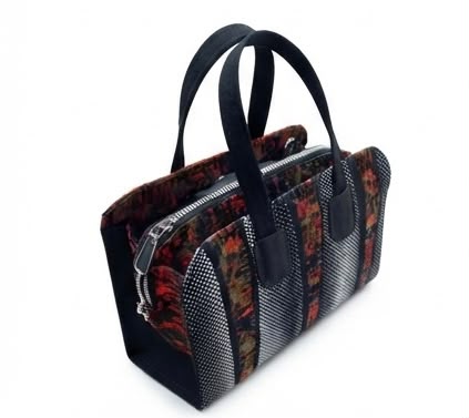 Side angle view of Ankara patchwork bowling bag with alternating black and white polka dot and dark floral print panels, black top handles and silver zip